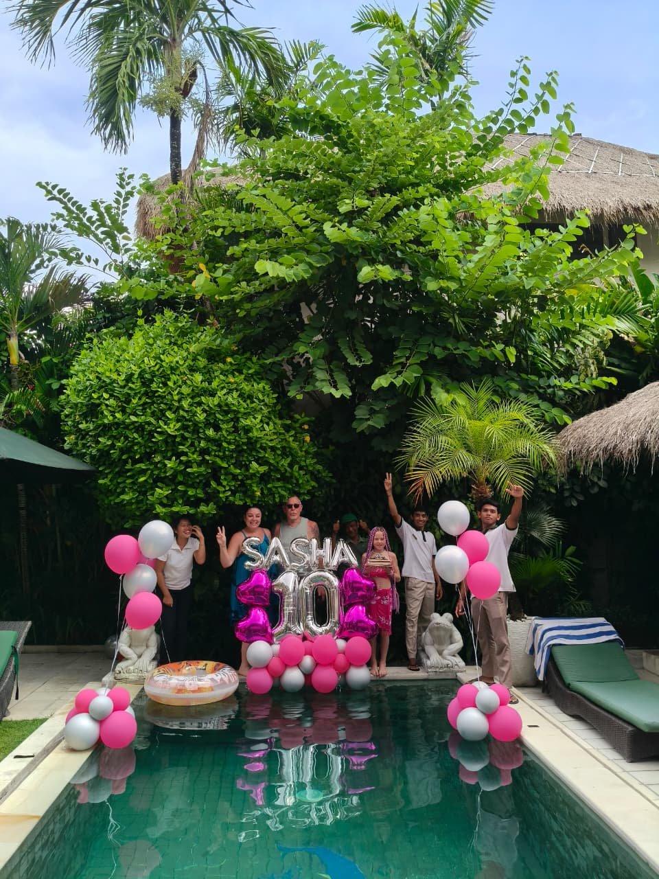 A 10th birthday celebration by the pool at Villa Krisna, featuring a family and villa staff posing with pink and silver balloon decorations spelling out the name Sasha and the number 10.