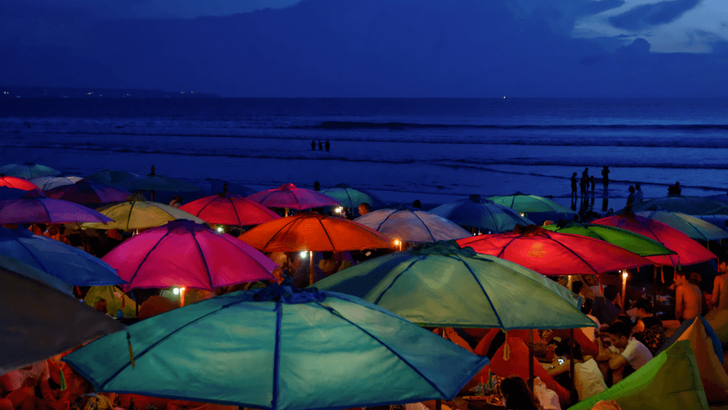 Evening scene at a Bali beach club with vibrant, colorful umbrellas lit by soft lights, overlooking the calm ocean waves under a deep blue twilight sky, creating a lively yet serene atmosphere for beachgoers.