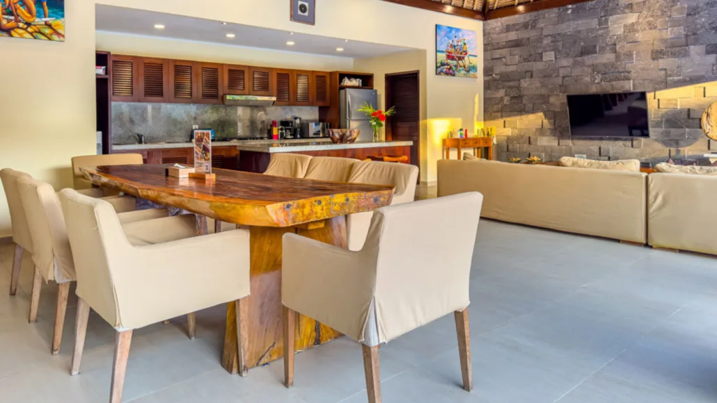 Spacious dining and living area in Villa Bugis, featuring a large rustic wooden dining table with cushioned chairs, a modern kitchen in the background, and a cozy lounge with stone-accented walls and a mounted flat-screen TV.