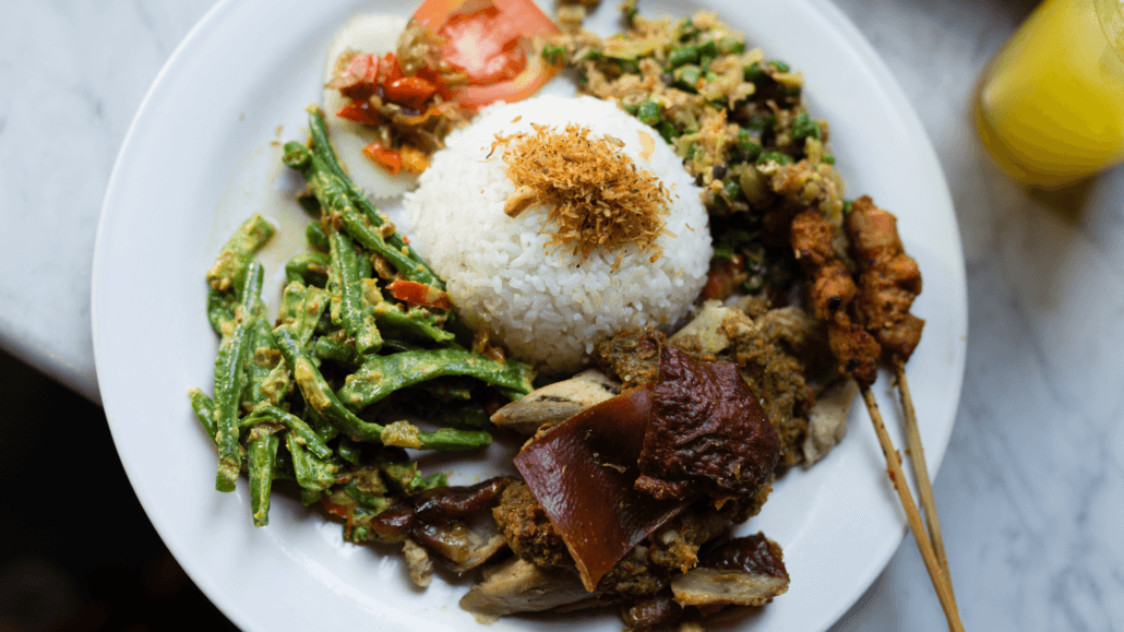 Traditional Balinese Babi Guling or roast pork with spiced shredded coconut, green bean salad, satay skewers, and assorted vegetables served on a white plate