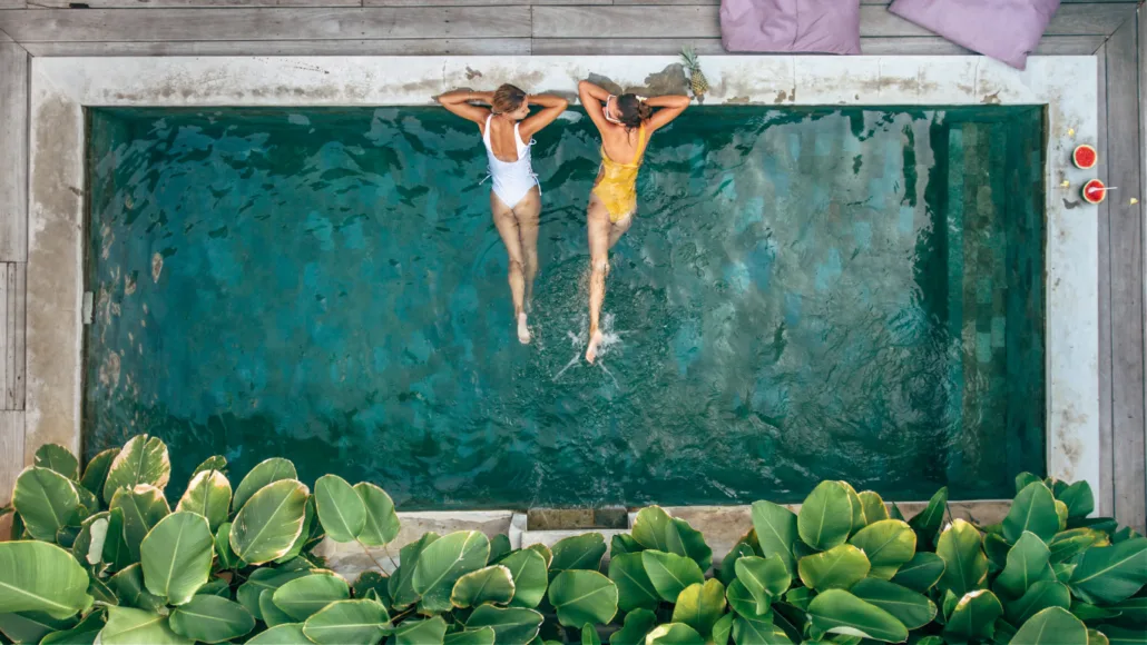 Top-down view of two women relaxing at the edge of a private pool at Villa Bugis, surrounded by lush green plants, with refreshing drinks and vibrant decor nearby, creating a tranquil and luxurious holiday atmosphere.
