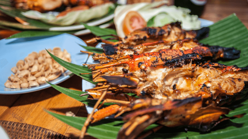 Grilled Balinese seafood featuring charred prawns served on banana leaves with a side of peanuts, fresh vegetables, and lime slices on a wooden table.