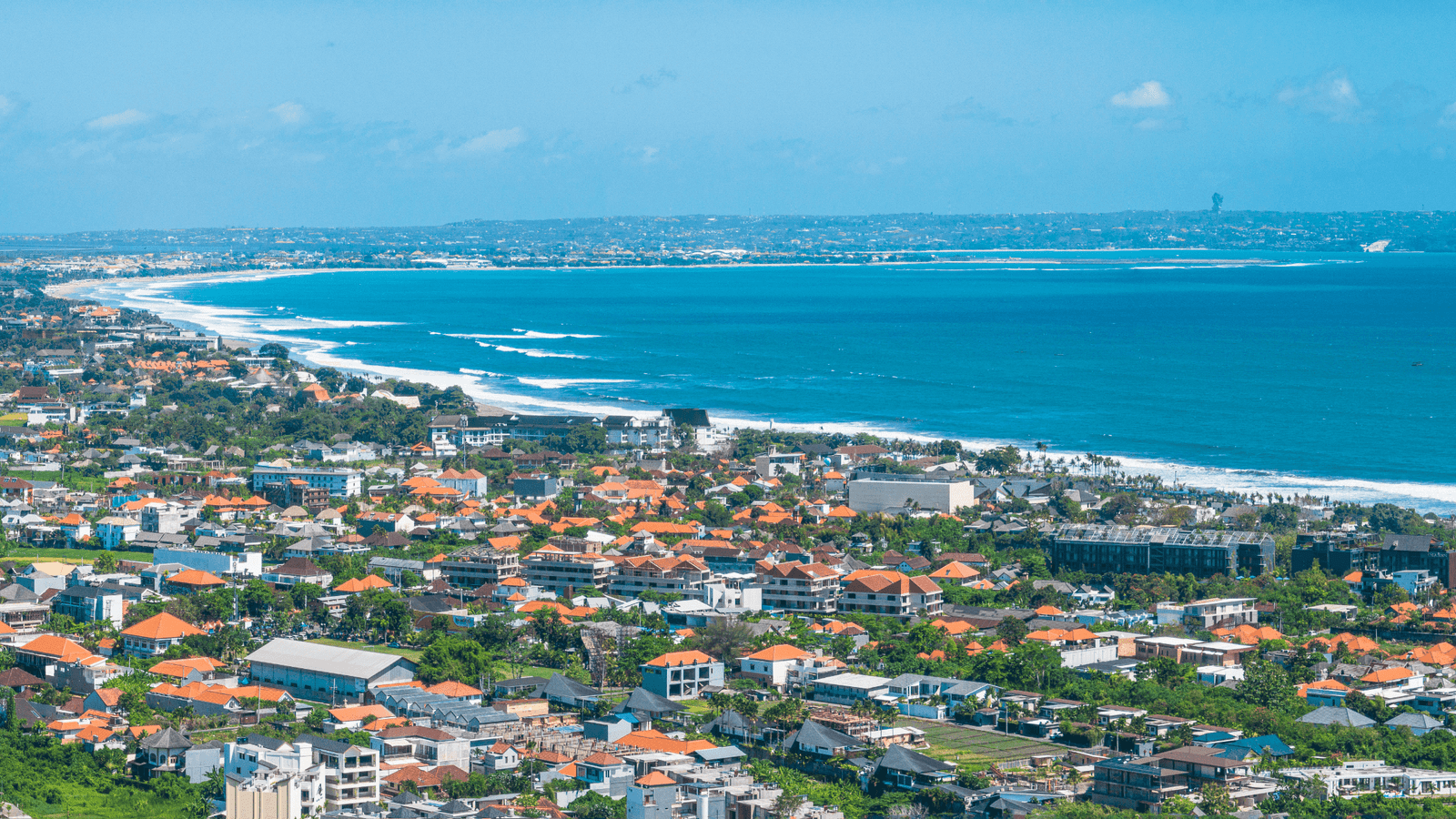 A picturesque aerial view of Bali's coastal town with orange-roofed buildings, vibrant greenery, and a stunning blue ocean stretching into the horizon.
