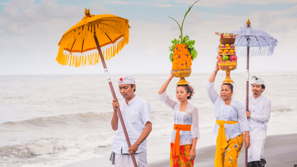 A group of Balinese people dressed in traditional attire carrying colorful ceremonial offerings under decorative umbrellas, walking along the beach against a backdrop of the sea.
