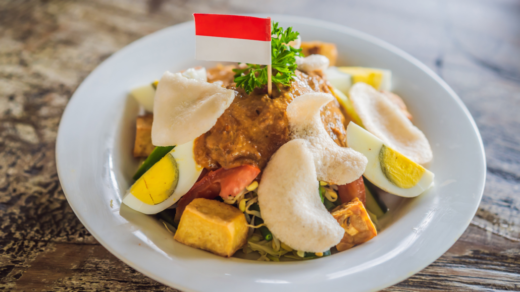 Traditional Indonesian Gado-Gado salad with peanut sauce, boiled eggs, tofu, vegetables, and prawn crackers, garnished with an Indonesian flag on a white plate.