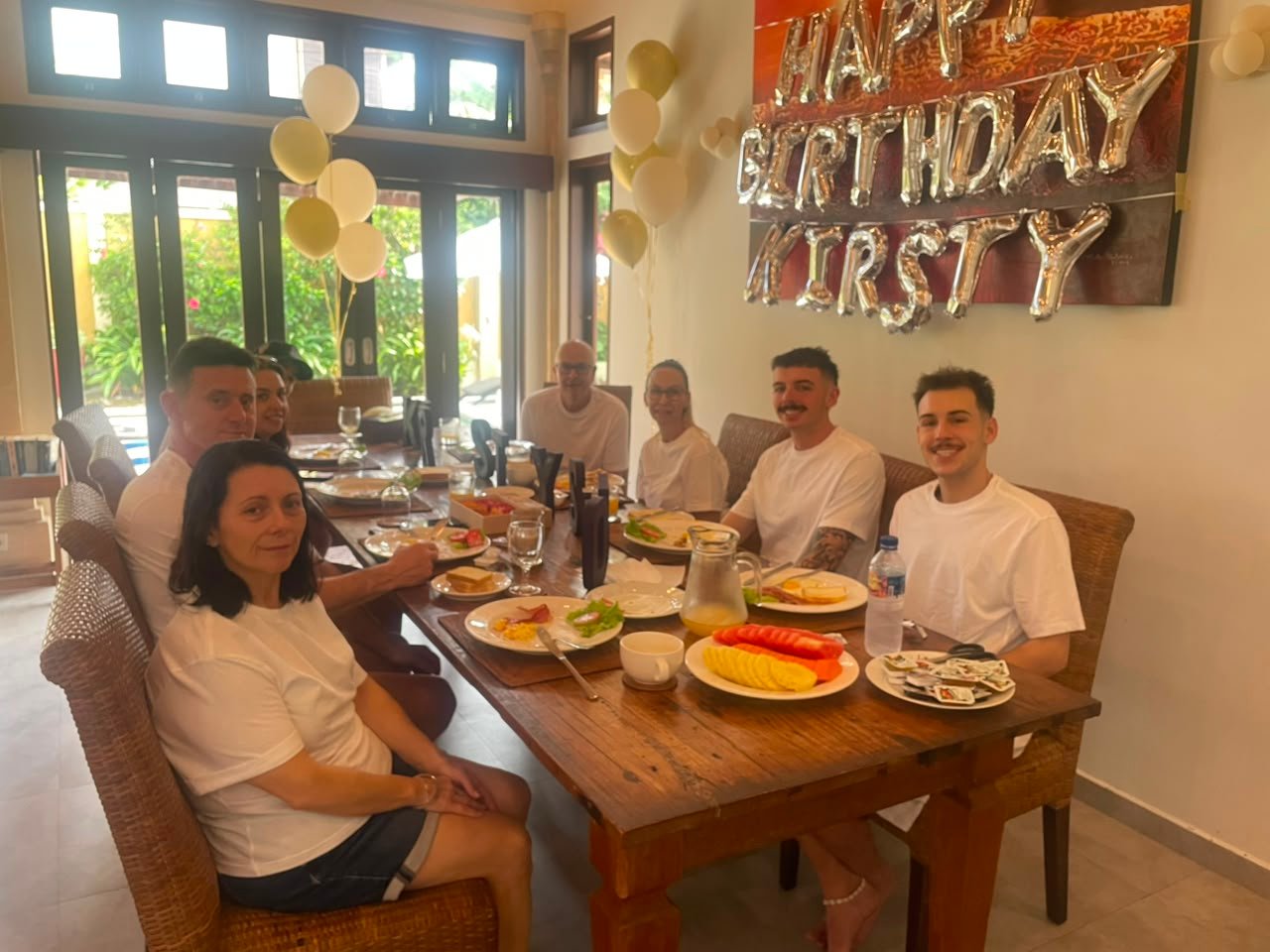 A group of family and friends gathering for a birthday breakfast around a large dining table at Villa Gembira, decorated with Happy Birthday Kirsty balloons.