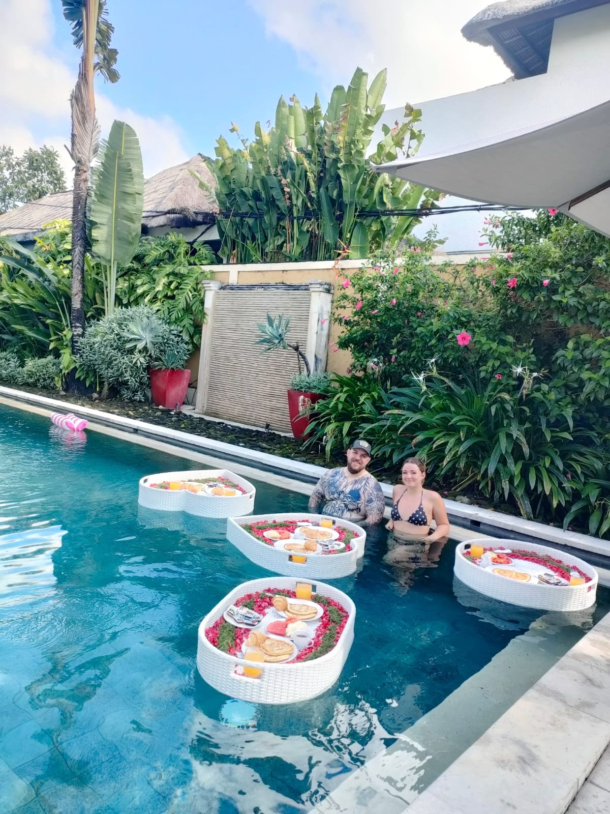 A couple relaxing in a private swimming pool at Villa Gembira surrounded by three heart-shaped floating breakfast trays filled with tropical fruits and pastries.