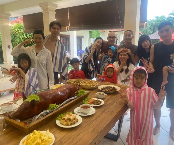 A large family with young children wrapped in pool towels posing happily behind a dining table set with a traditional Balinese roasted pig, Babi Guling, at Villa Vara.