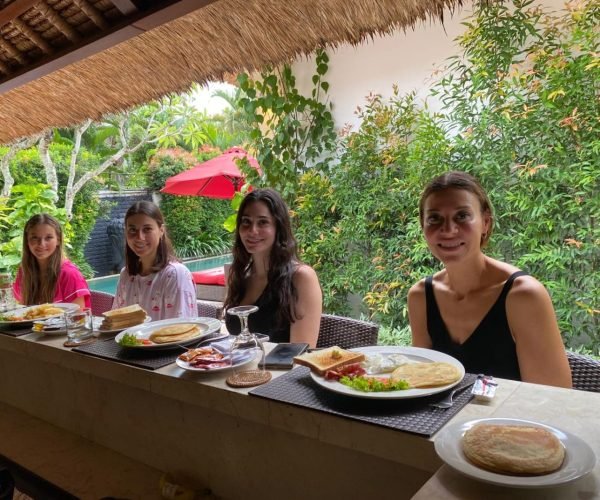 Four female guests smiling and enjoying a delicious morning breakfast at the outdoor kitchen bar counter surrounded by lush greenery at Villa Jaclan.