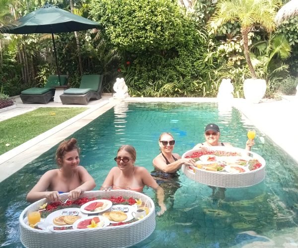 Three female guests relaxing in the private swimming pool while enjoying a delicious floating breakfast experience on woven trays at Villa Krisna.