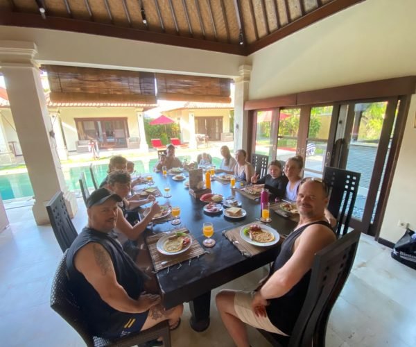 A large family and friends group smiling while enjoying a fresh morning breakfast spread at a long dark wooden dining table overlooking the pool at Villa Santi.