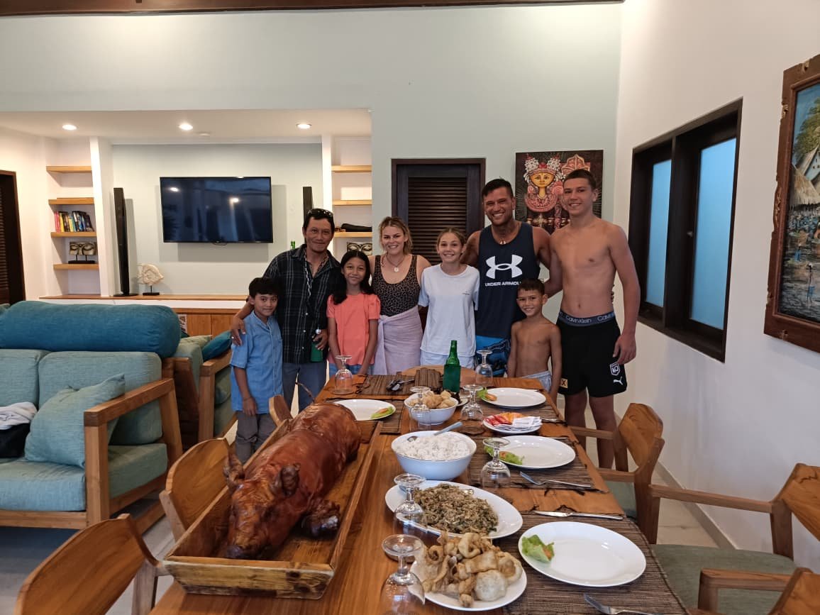 A family group posing happily behind a long wooden dining table set with a traditional Balinese roasted pig feast, known as Babi Guling, inside Villa Rama.