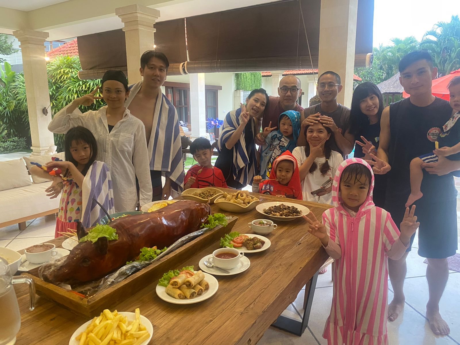 A large family with young children wrapped in pool towels posing happily behind a dining table set with a traditional Balinese roasted pig, Babi Guling, at Villa Vara.