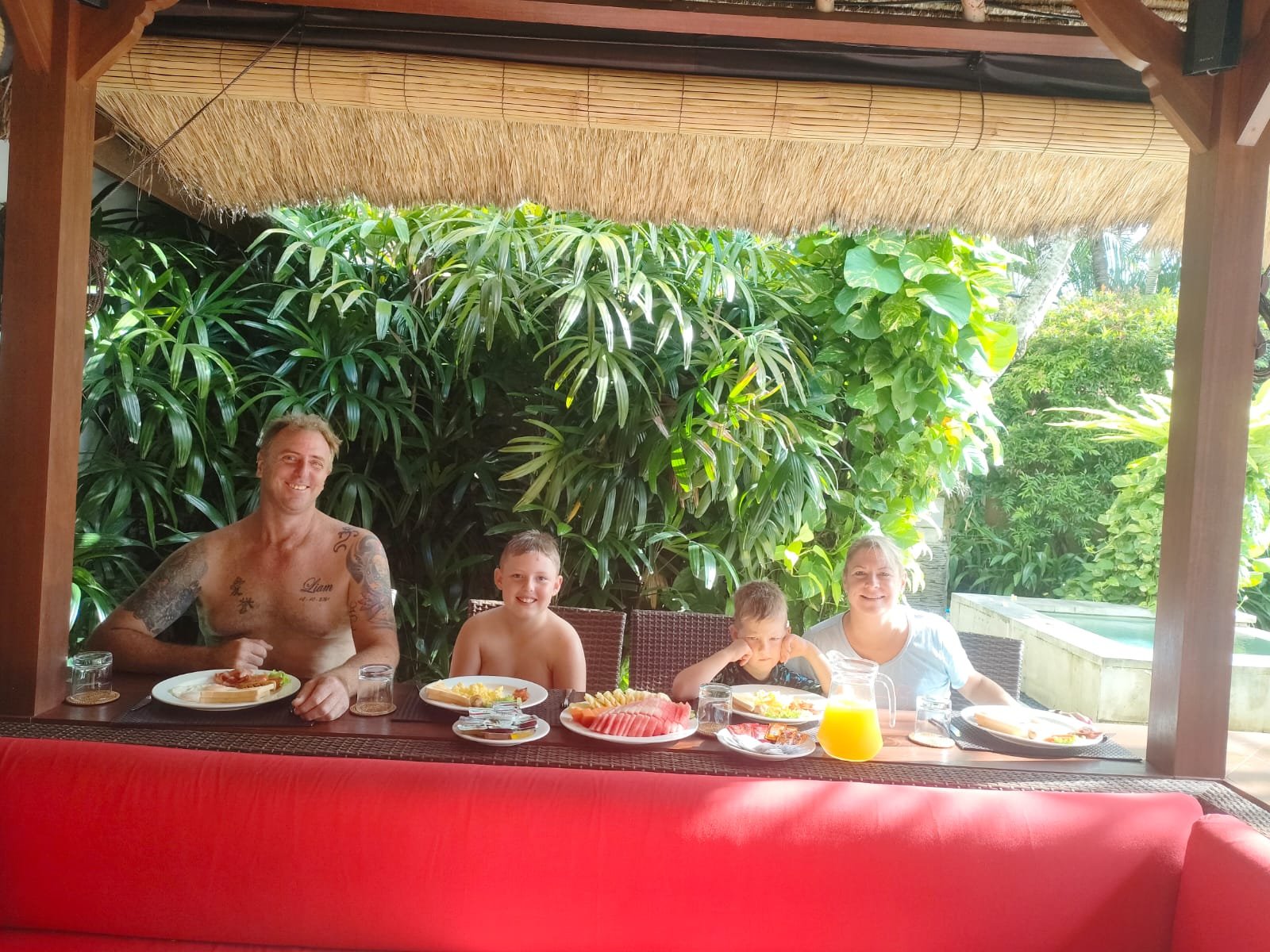 A family with two young boys enjoying a morning breakfast spread together inside a traditional thatched-roof gazebo surrounded by tropical plants at Villa Jaclan.