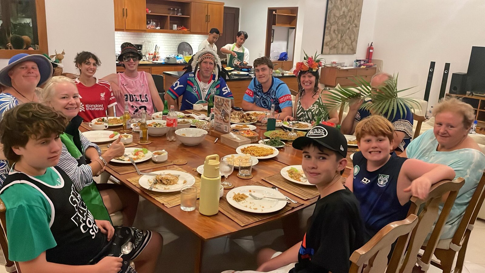 A multi-generational family enjoying a fun, catered dinner gathering at a large table while a private chef cooks in the background at Villa Alore.