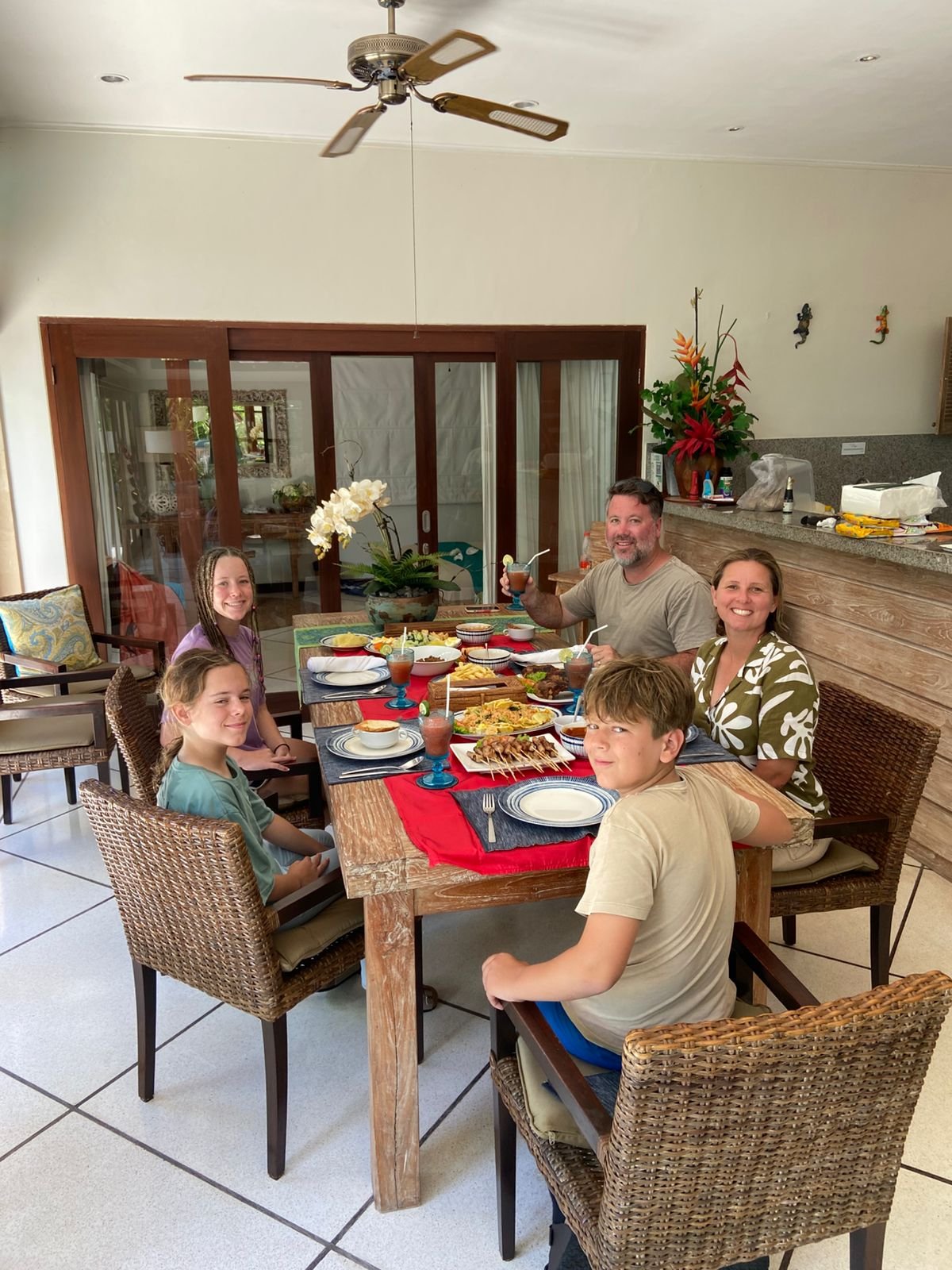 A smiling family of five sitting around a large wooden dining table, ready to enjoy a freshly prepared traditional Indonesian meal together inside Villa Nakula.