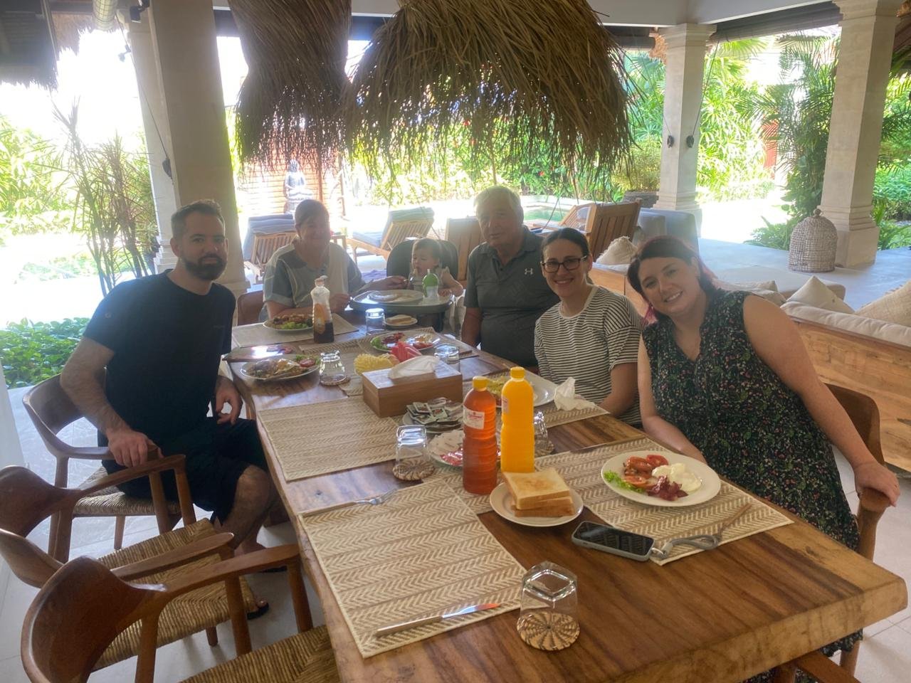 A multi-generational family enjoying a casual lunch together at a large wooden dining table in the bright, open-air living area of Villa Olive in Seminyak.
