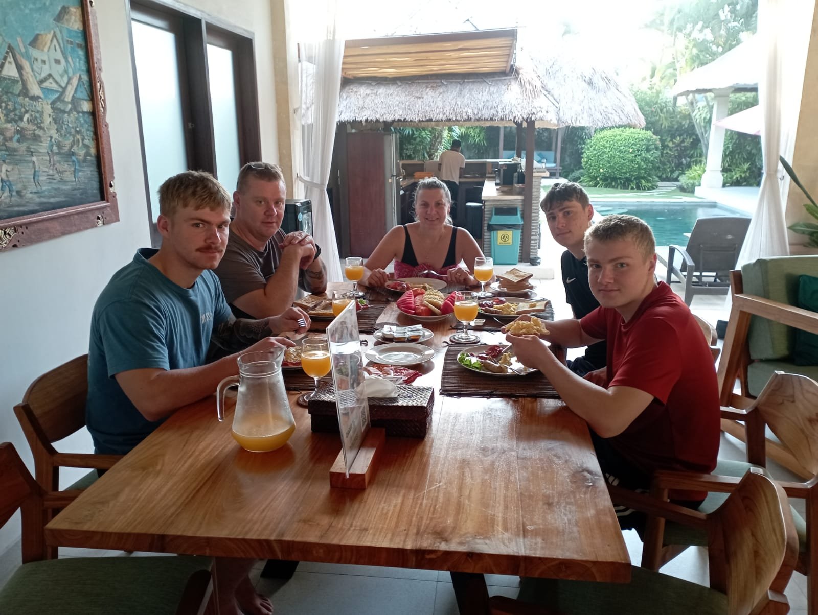 A family of five enjoying a fresh morning breakfast together at a large wooden dining table with a beautiful view of the swimming pool at Villa Rama.