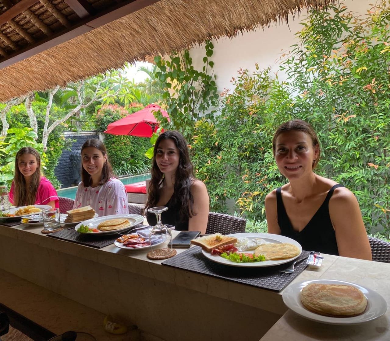 Four female guests smiling and enjoying a delicious morning breakfast at the outdoor kitchen bar counter surrounded by lush greenery at Villa Jaclan.