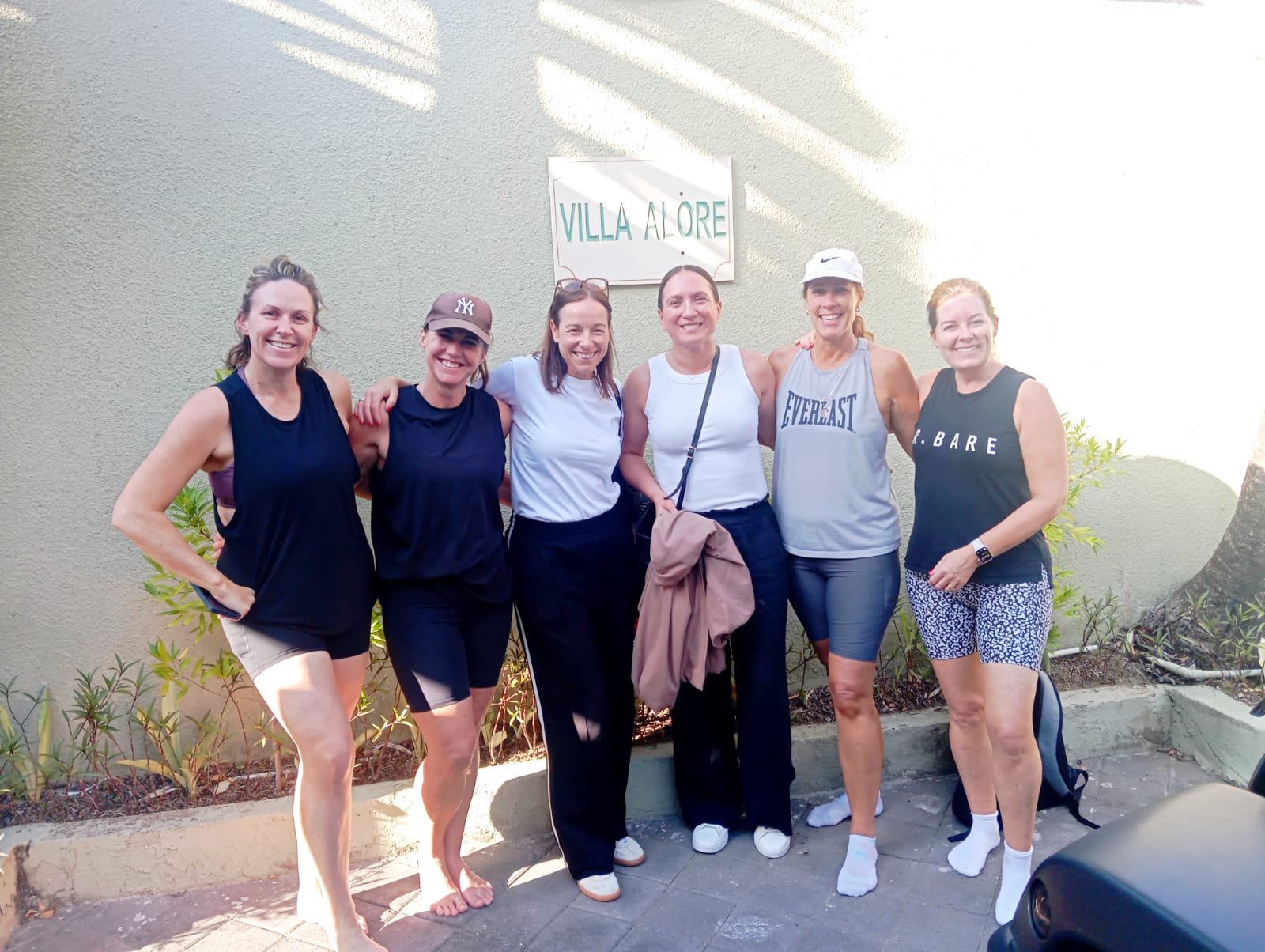 Six happy female guests in casual activewear posing together in front of the Villa Alore entrance sign.