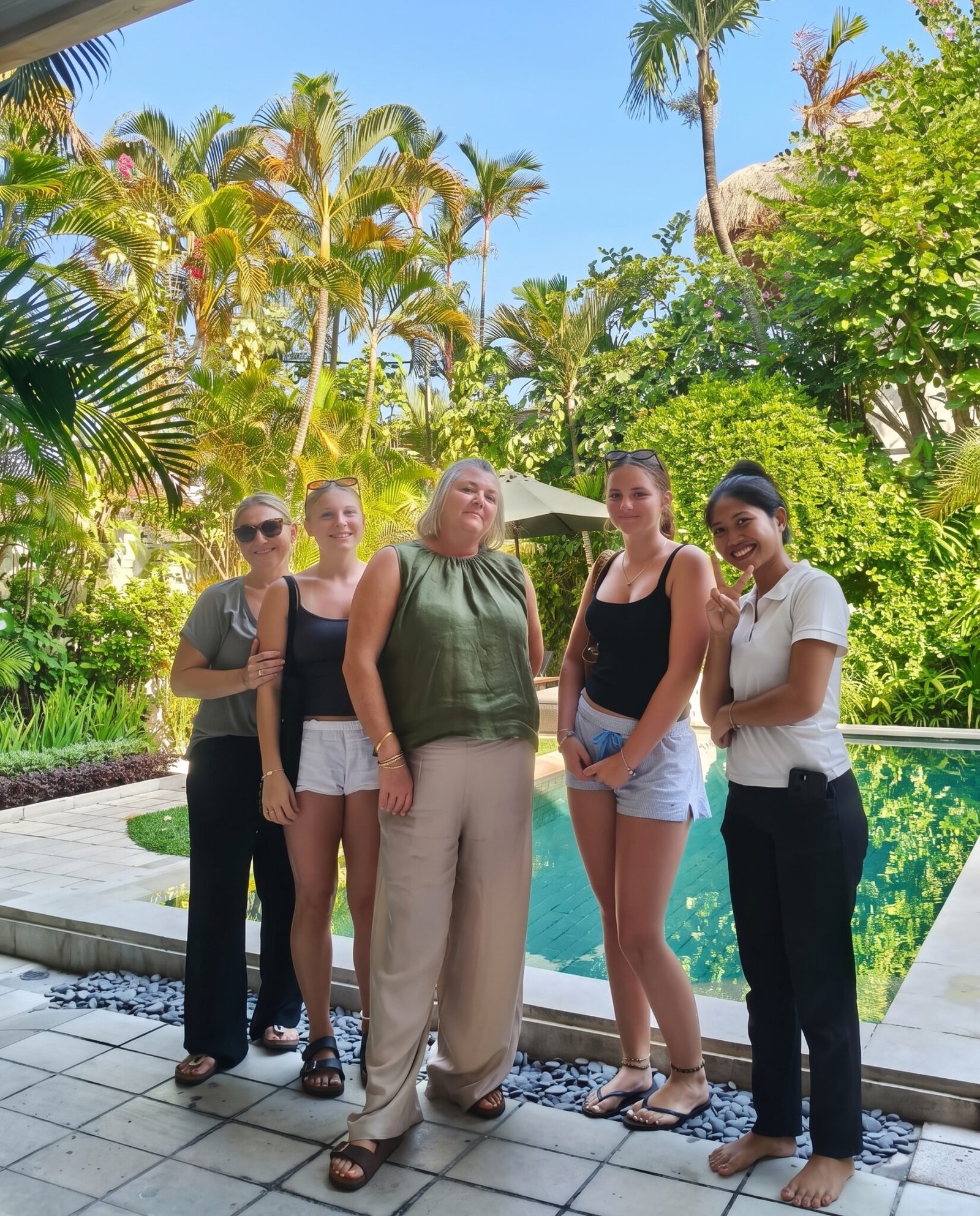 Four female guests smiling and posing for a group photo with a friendly villa staff member next to the private swimming pool surrounded by tropical gardens at Villa Krisna in Seminyak.