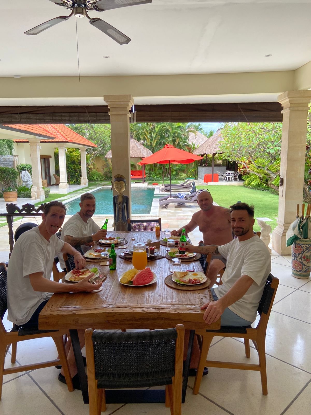 Four male guests smiling and raising Bintang beers while enjoying a fresh breakfast spread at a wooden dining table with a view of the garden and pool at Villa Vara.