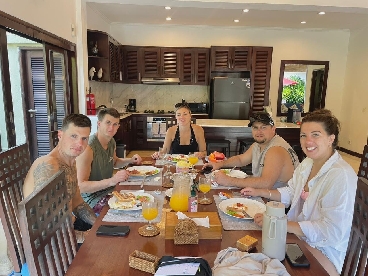 A group of five friends gathering for a morning breakfast around a large wooden table near the kitchen area at Villa Kebun in Seminyak.