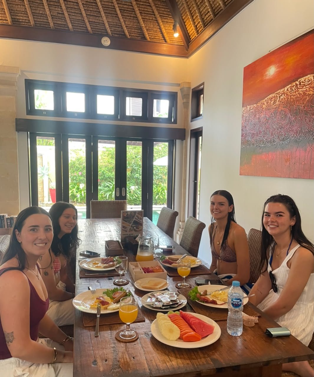 Four young women smiling while enjoying a morning breakfast together at a large wooden dining table inside Villa Gembira.