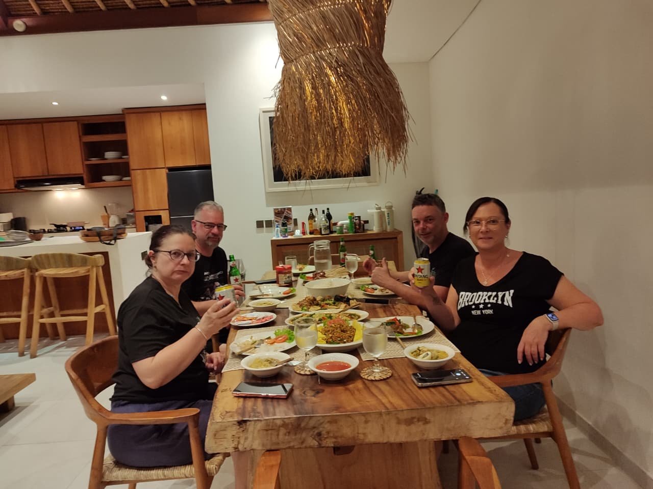 Four adult friends enjoying a delicious dinner spread and Bintang beers at a beautiful wooden dining table next to the kitchen inside Villa Olive.