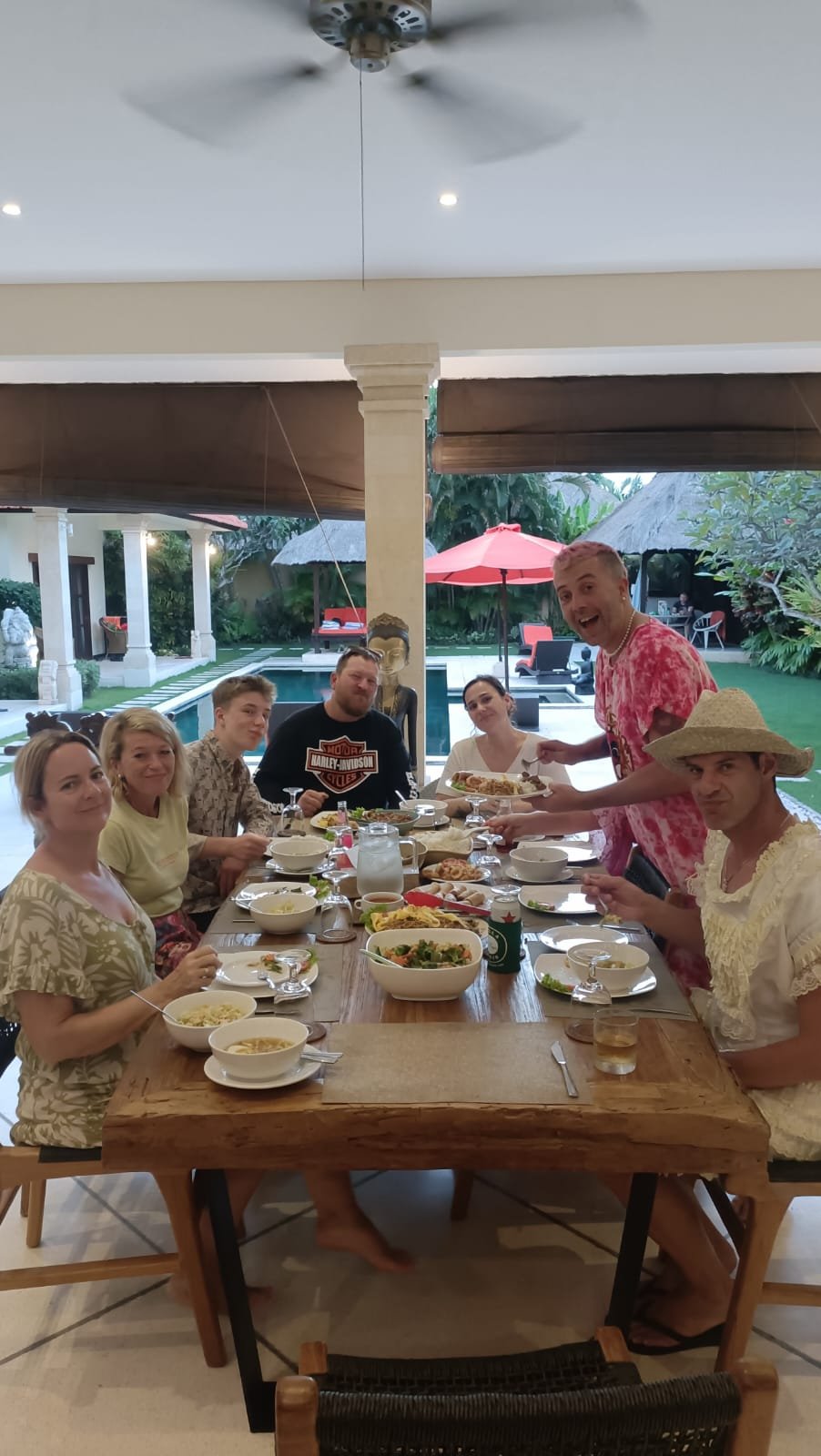A group of friends enjoying a casual dinner gathering at a large wooden dining table in the open-air living space overlooking the swimming pool at Villa Vara in Seminyak.