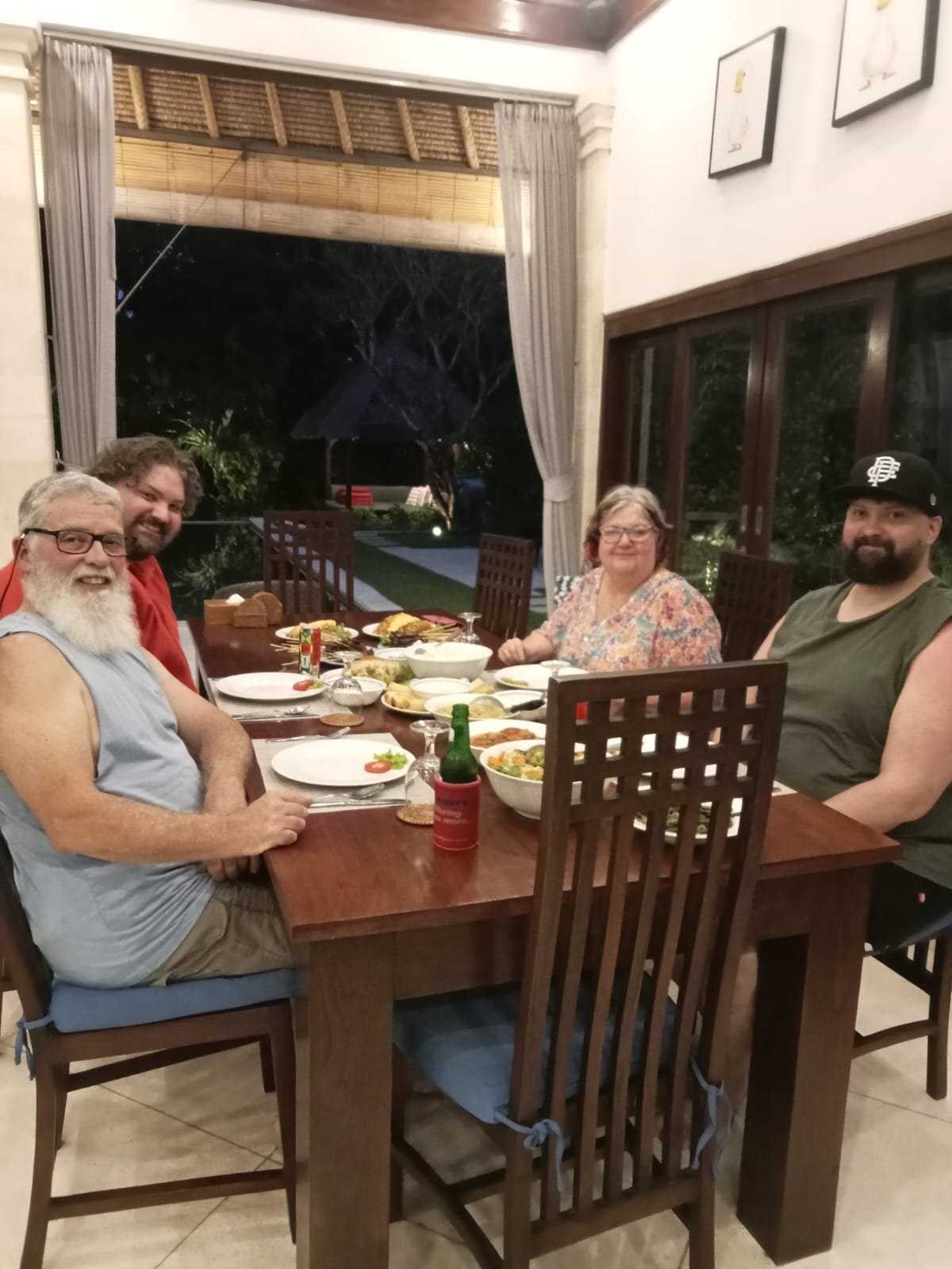 Four guests enjoying an evening dinner together at a wooden dining table in the open-air living space of Villa Kebun in Seminyak.