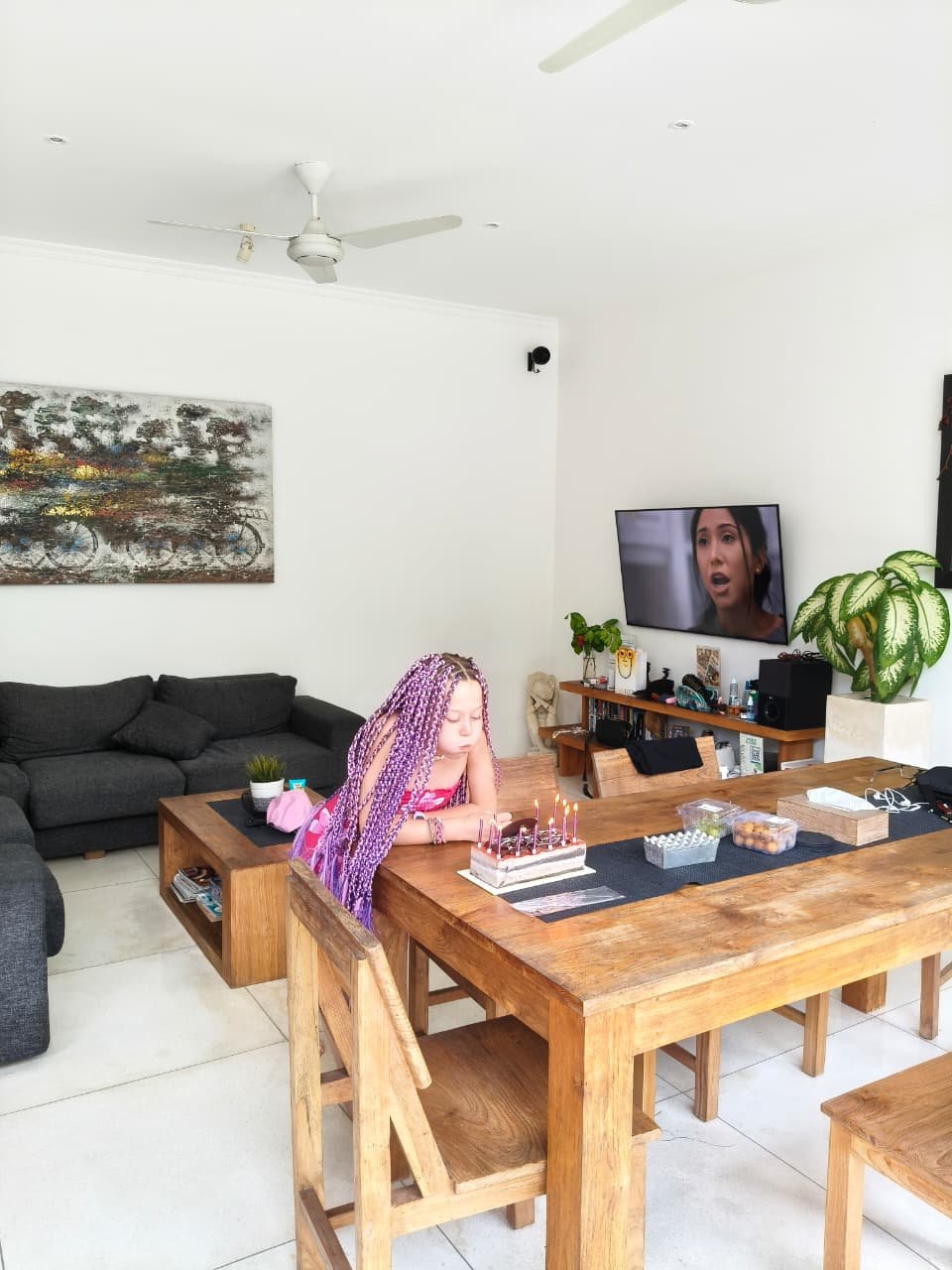 A young girl with purple braided hair leaning over a wooden dining table to blow out the candles on her chocolate birthday cake inside the open living area of Villa Krisna.