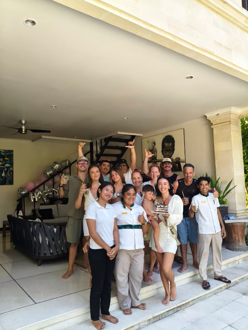 A large group of guests and three friendly villa staff members posing for a photo during a birthday celebration, featuring a woman wearing a Birthday Queen sash holding a cake at Villa Nakula.