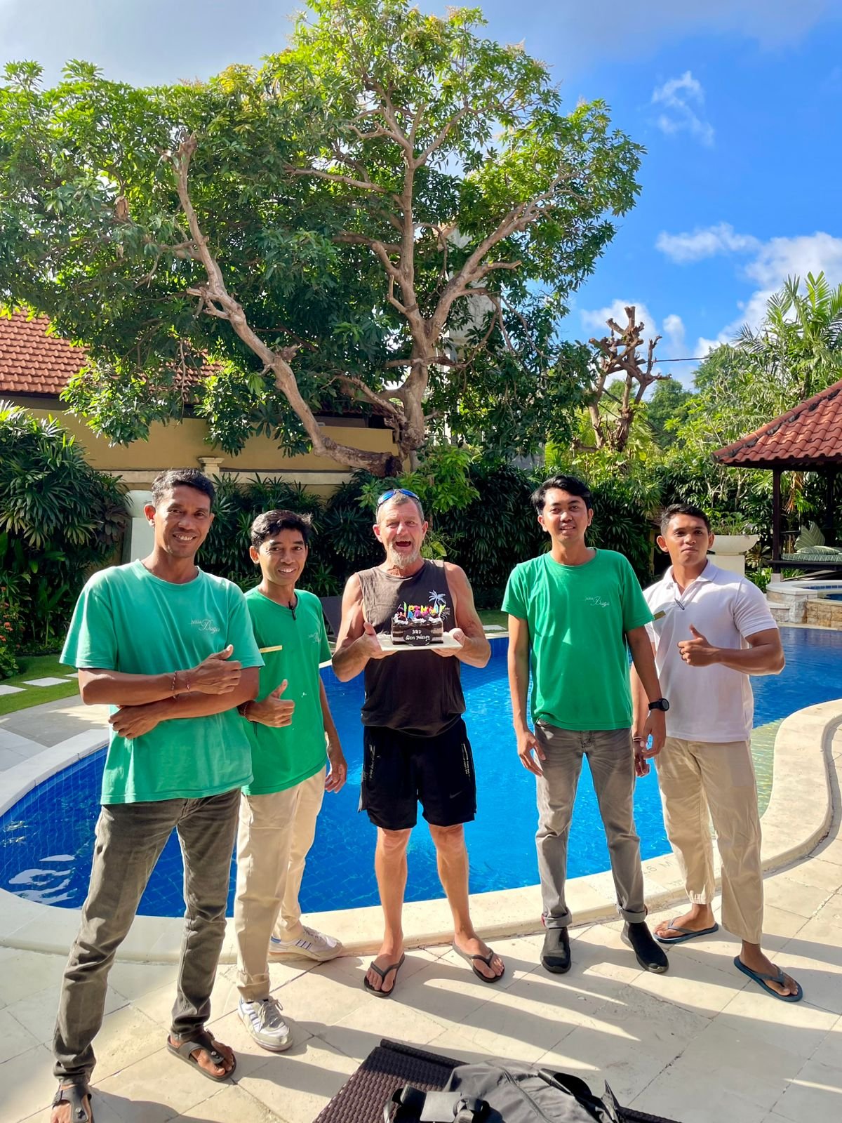A senior male guest holding a birthday cake, smiling and posing with three villa staff members by the private pool at Villa Ginger in Seminyak.