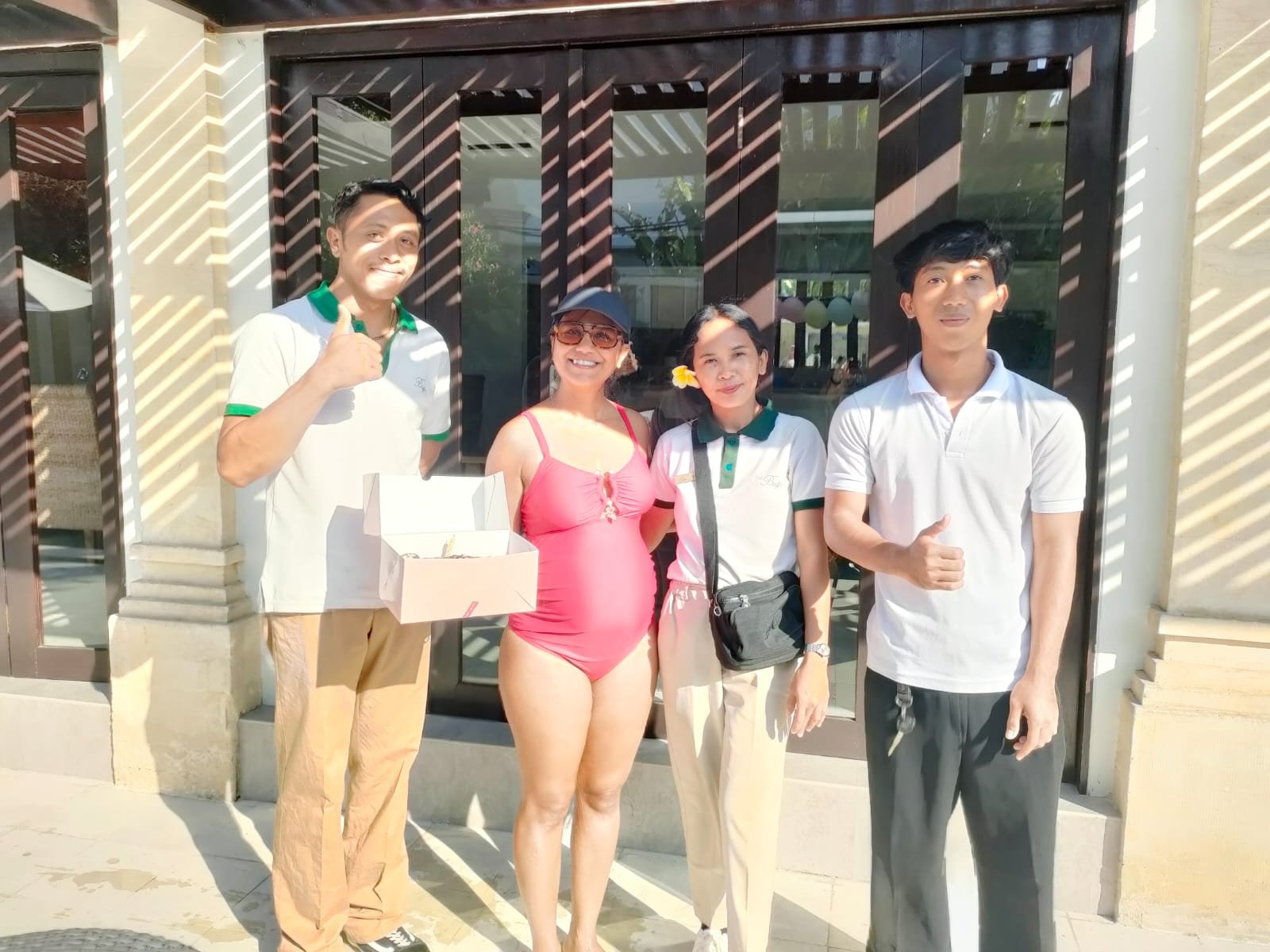 A female guest in a pink swimsuit holding a birthday cake box, posing with three friendly villa staff members outside Villa Gembira in Seminyak.