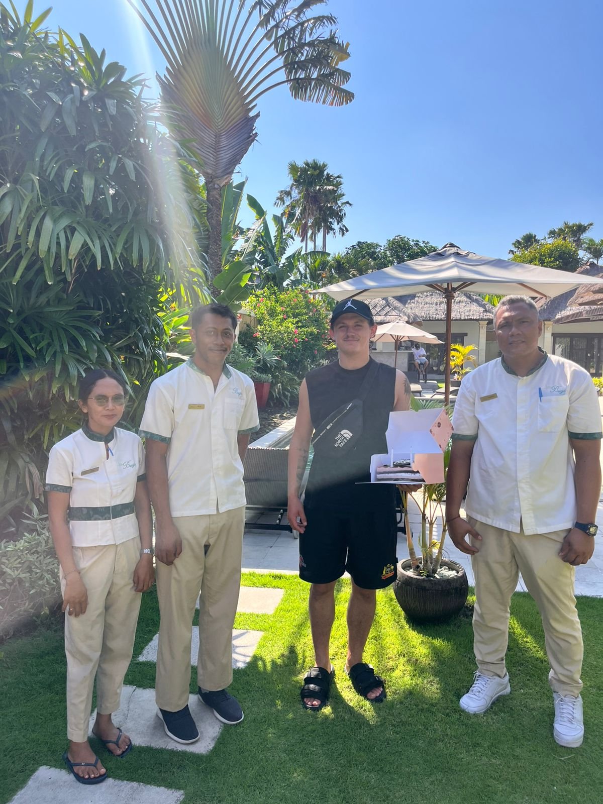 A male guest holding a birthday cake box standing alongside three smiling villa staff members in the lush tropical garden of Villa Gembira.