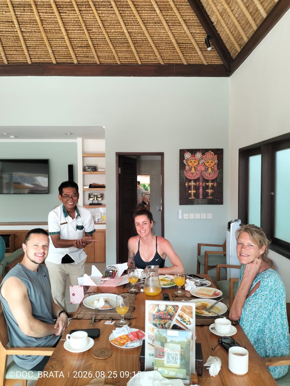 A smiling villa staff member presenting a birthday cake to three guests sitting at a wooden dining table during breakfast inside Villa Rama.