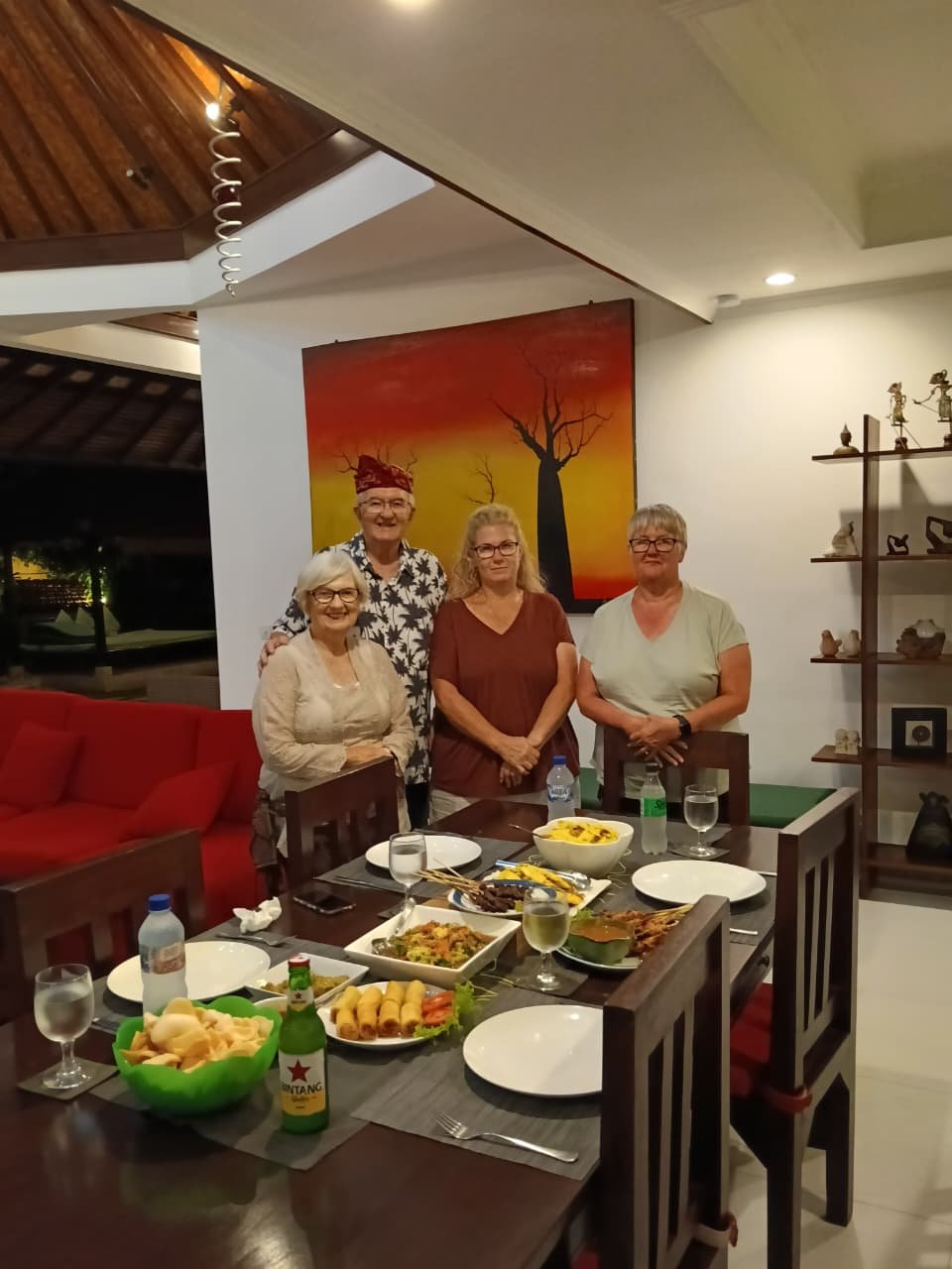 Four senior guests standing behind a dining table filled with a traditional Balinese dinner spread, including satay and Bintang beer, inside Villa Ginger.