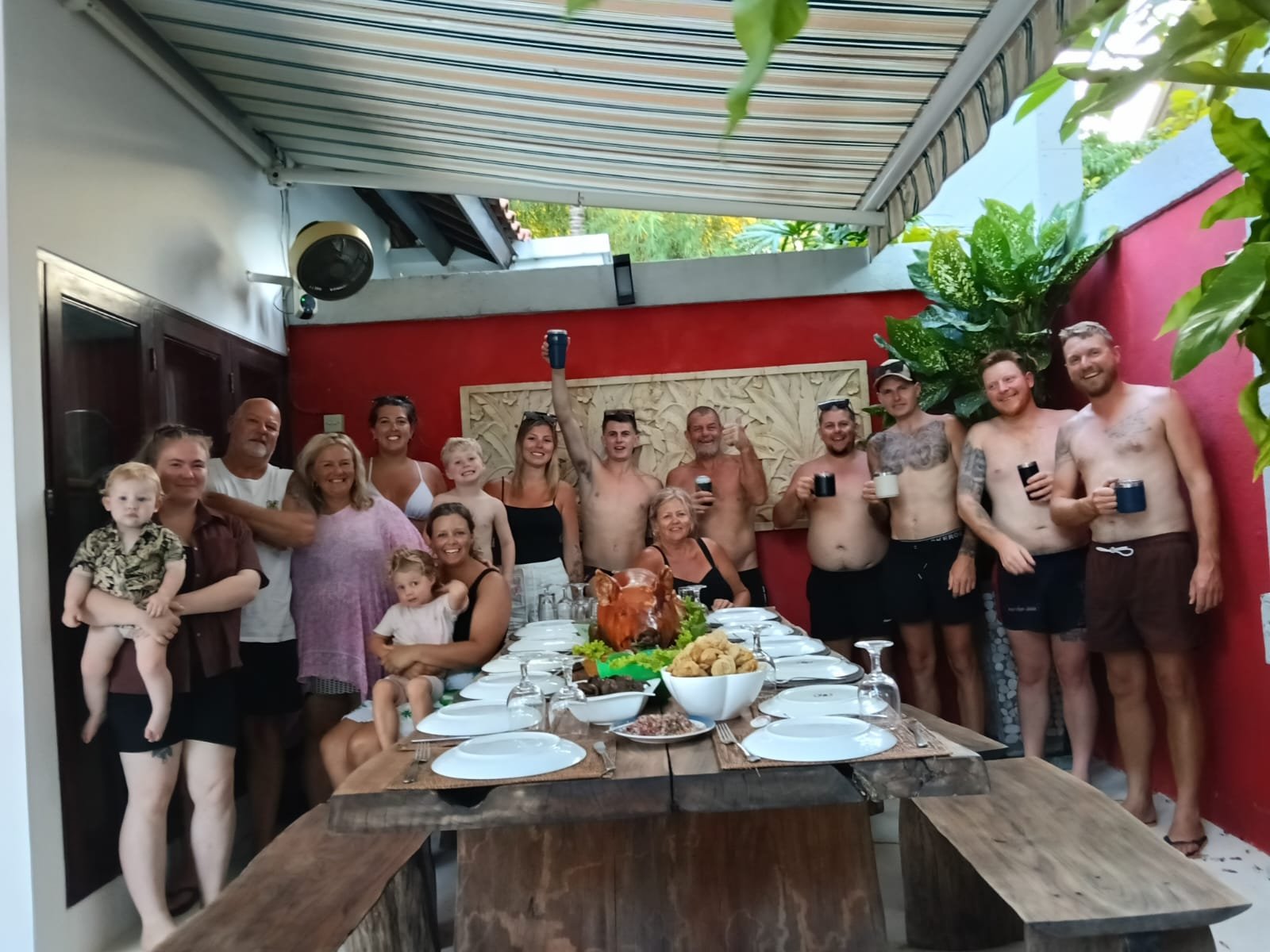 A large group of family and friends gathering for a traditional Balinese roasted pig feast at a long wooden dining table in the outdoor patio of Villa Ginger.
