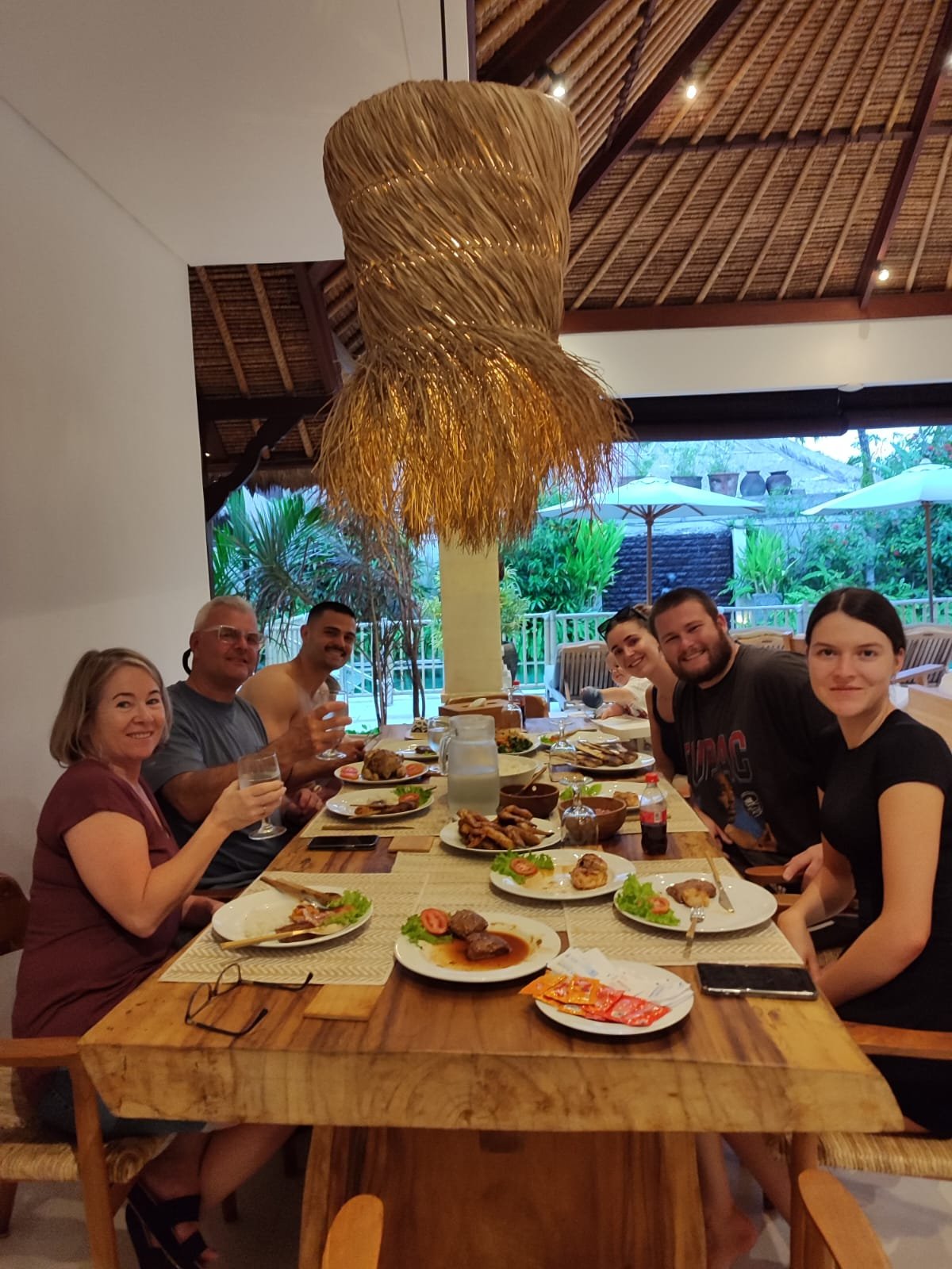 A group of family and friends smiling and raising their glasses during an evening dinner gathering at a long wooden table under the thatched roof of Villa Olive.