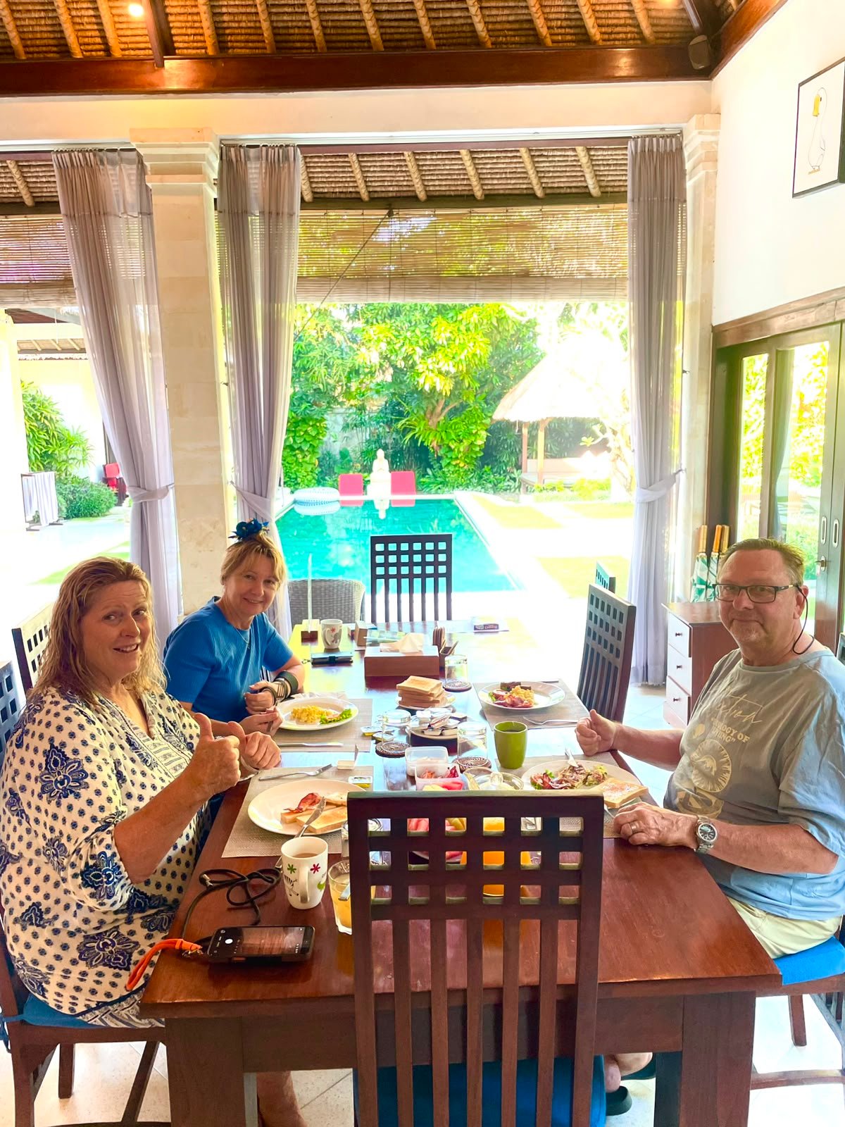 Three guests smiling and giving thumbs up while eating a fresh morning breakfast at the dining table with a beautiful view of the pool at Villa Kebun.