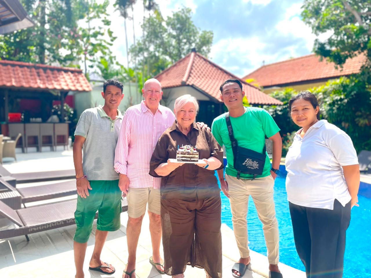 An older couple celebrating a birthday with a cake, posing with two friendly villa staff members next to the swimming pool at Villa Ginger in Seminyak.