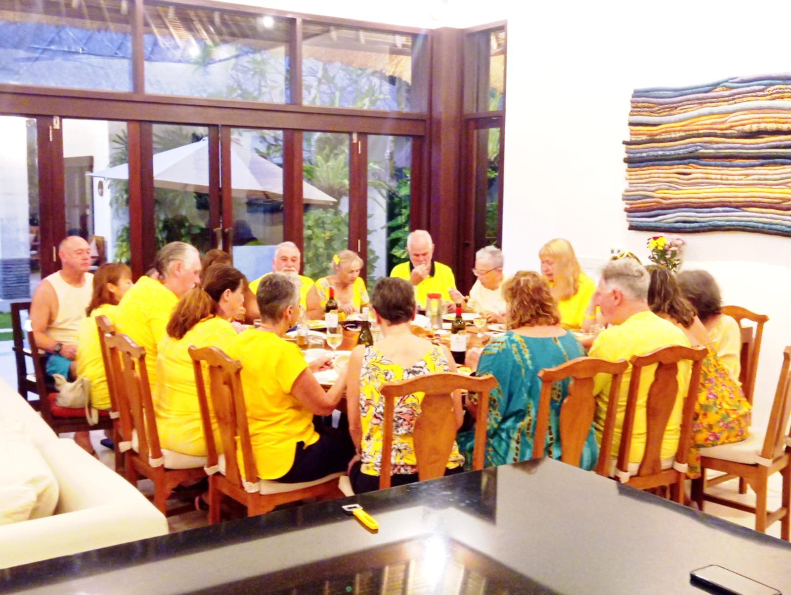 A large group of senior guests in matching yellow shirts enjoying a group dinner at a long wooden table inside Villa Alore in Seminyak.