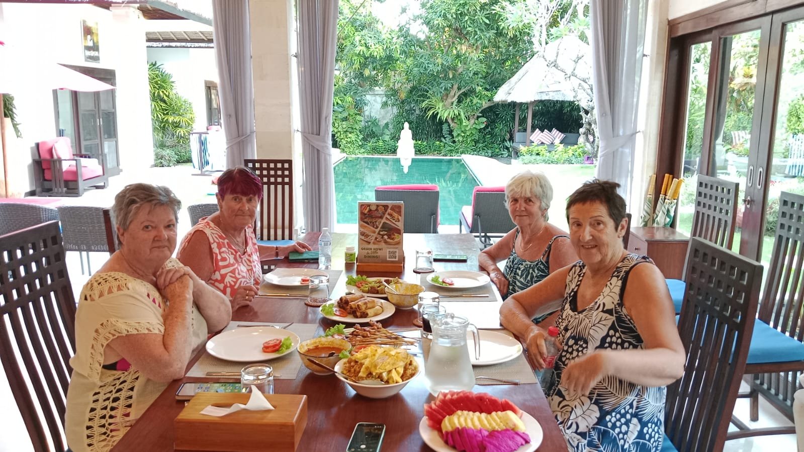Four senior female guests smiling while enjoying a delicious lunch with satay and fresh fruit at a dining table overlooking the pool at Villa Kebun.