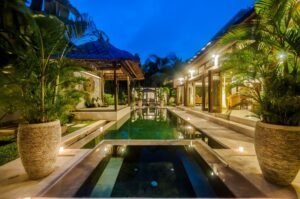 Private pool lit at dusk with stone planters and tropical palms at Villa Damai Seminyak