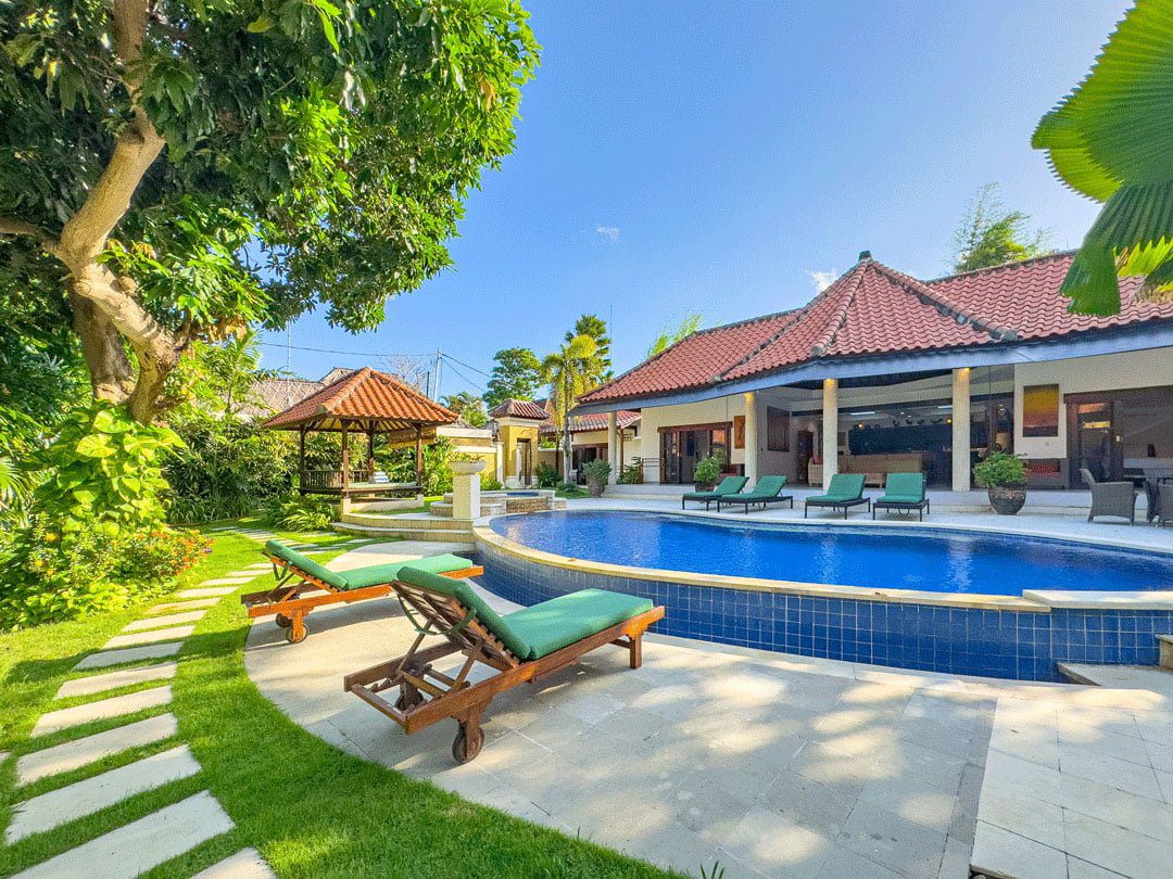 Bright daytime view of the main poolside deck at Villa Ginger Seminyak by Villa Bugis Seminyak villas, showcasing a clear blue pool surrounded by lush greenery.