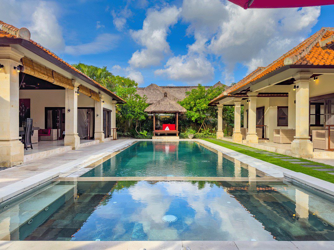 Bright daytime view of the main poolside deck at Villa Santi Seminyak by Villa Bugis Seminyak villas, showcasing a clear blue pool surrounded by exotic greenery.