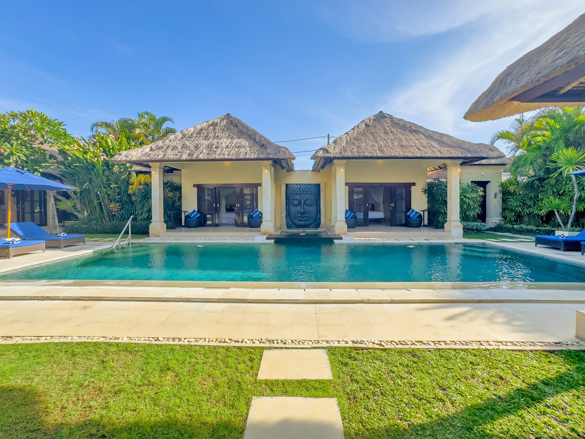 Bright daytime view of the main poolside deck at Villa Saphir Seminyak by Villa Bugis Seminyak villas, showcasing a clear blue pool surrounded by exotic greenery.