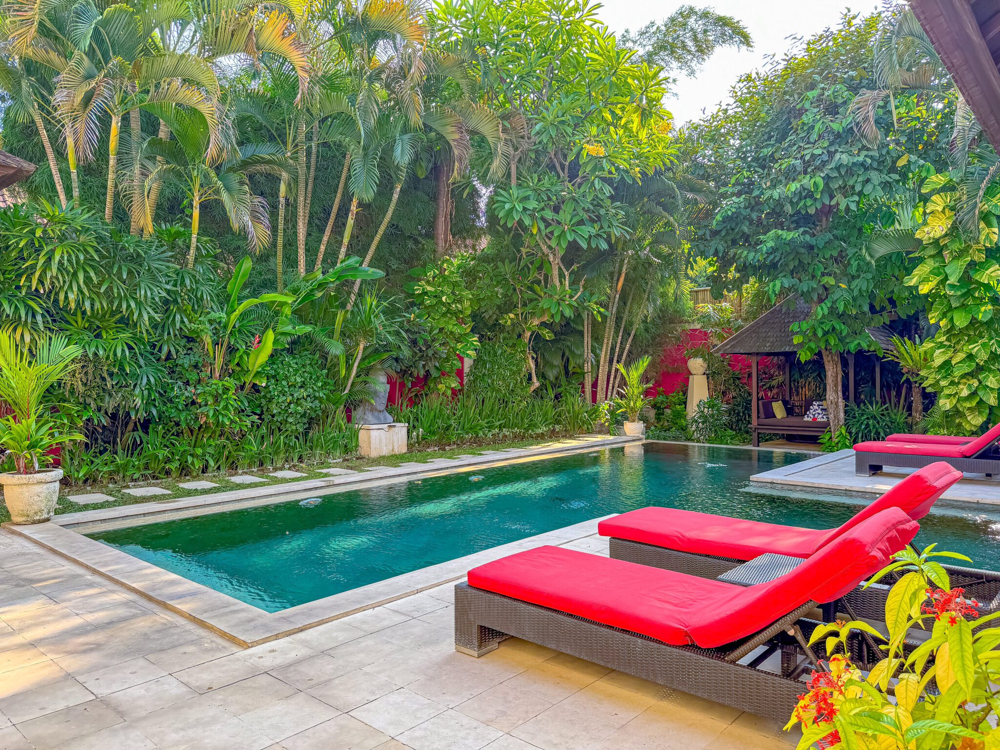 Bright daytime view of the main poolside deck at Villa Tresna Seminyak by Villa Bugis Seminyak villas, showcasing a clear blue pool surrounded by exotic greenery.