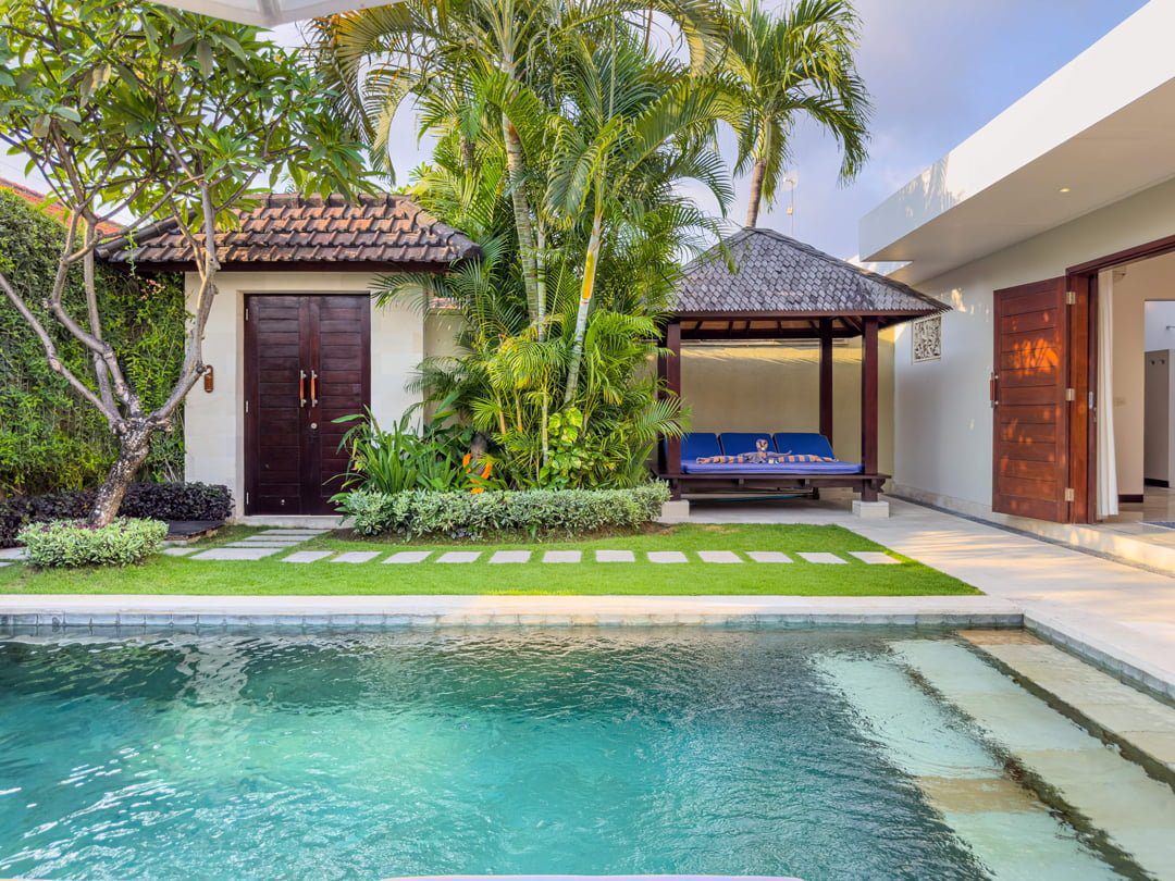 Bright daytime view of the main poolside deck at Villa Umah Kupu-Kupu 2 Seminyak by Villa Bugis Seminyak villas, showcasing a clear blue pool surrounded by exotic greenery.